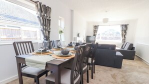 A dining area with a table set for six, surrounded by dark wooden chairs with light-colored cushions. A dark sofa and a television are visible in the background. - 38 Clive Avenue (Lytham St. Anne's)