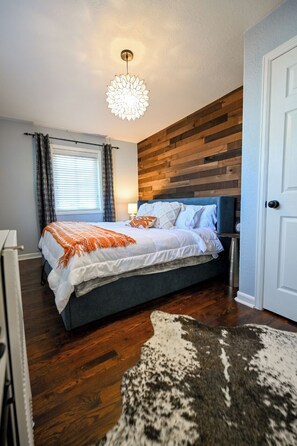 Un dormitorio con cama, cabecera de madera, ventana con cortinas, mesita de noche y alfombra.