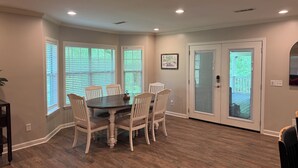 A dining room with a round table and six chairs, large windows with blinds, a door with a glass panel, and recessed ceiling lighting.