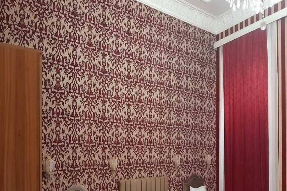 Ein Hotelzimmer mit drei hölzernen Betten, einem gemusterten Wandteppich, einem roten Vorhang und einer Kristallleuchter.