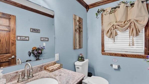 Un baño con lavamanos de mármol, tocador de madera, inodoro y ventana con cortinas.