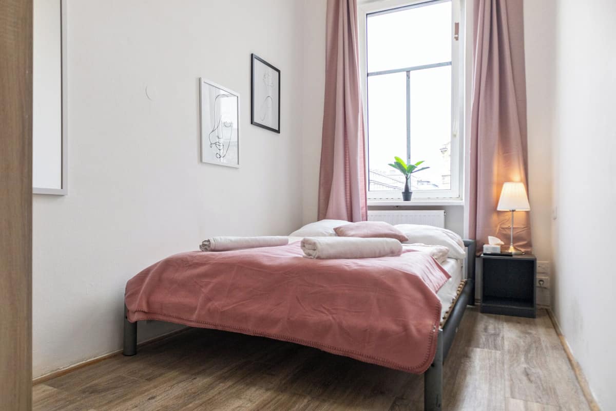 A neatly made bed with a pink blanket, a bedside lamp, and a window with curtains.
