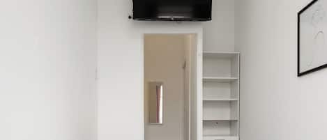 A bedroom with a bed, a TV mounted on the wall, a wardrobe, and a door leading to another room.