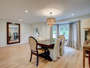 Comedor con mesa y sillas de madera, una lámpara de araña y ventanas grandes con cortinas.