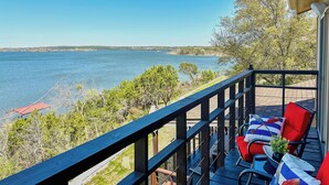 A wooden deck with red and blue striped cushions, a small table with a plant, and a view of a lake and distant shoreline. - Luxe Lake Granbury Retreat: Dock, Deck & Game Room (Granbury)