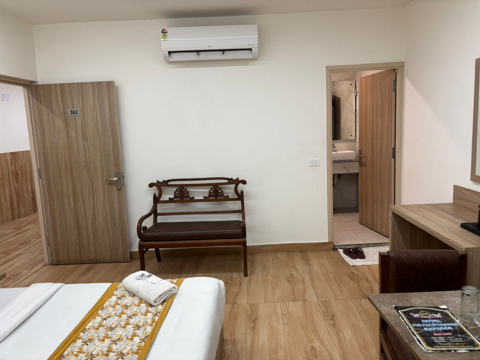 A hotel room with a bed, a wooden bench, a desk, and a bathroom visible through an open door.