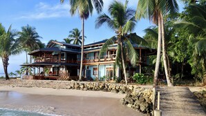 Une propriété située en bord de mer, dotée d’une terrasse en bois menant à un bâtiment à deux étages avec des balcons, le tout entouré de palmiers et bordé d’une plage de sable.