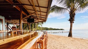 Un bar en bois avec des tabourets hauts, un palmier et une vue sur la plage.
