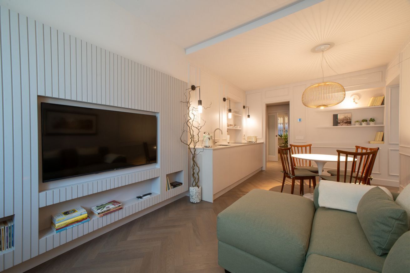 A modern living room with a flat-screen TV, a sofa, a dining area with a table and chairs, and a kitchen in the background.