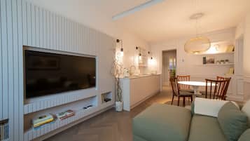 A modern living room with a flat-screen TV, a sofa, a dining area with a table and chairs, and a kitchen in the background.