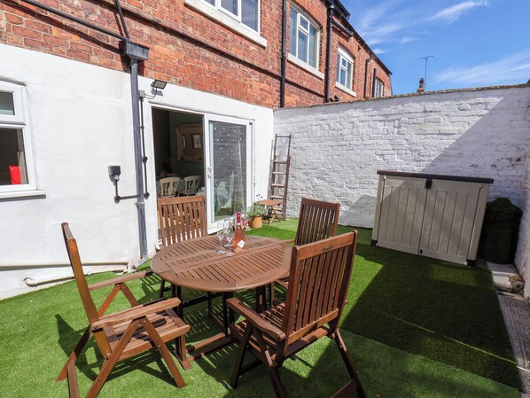 Une terrasse avec une table ronde en bois et des chaises, une porte blanche et un mur en briques.