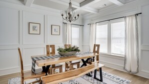 A dining area with a wooden table, benches, and a chandelier. There are framed pictures on the wall and a window with curtains.