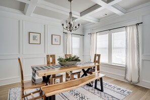 Un coin repas avec une table en bois, des bancs et un lustre. Des tableaux encadrés ornent le mur, et une fenêtre est agrémentée de rideaux.