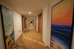 A hotel corridor with a patterned carpet, large paintings on the walls, and a long hallway leading to a door at the end.