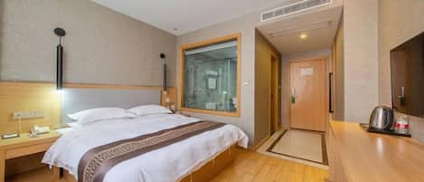 Quarto de hotel com cama, mesa de cabeceira e uma pequena escrivaninha com cadeira. Inclui área de chuveiro e cozinha compacta com micro-ondas e chaleira.