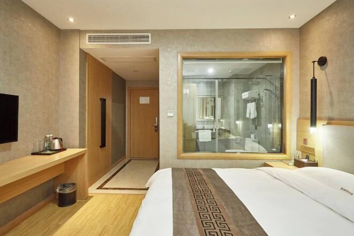 Habitació d'hotel amb un llit, una banca de fusta, un petit cubell de brossa, una finestra amb vista al bany i una televisió penjada a la paret.