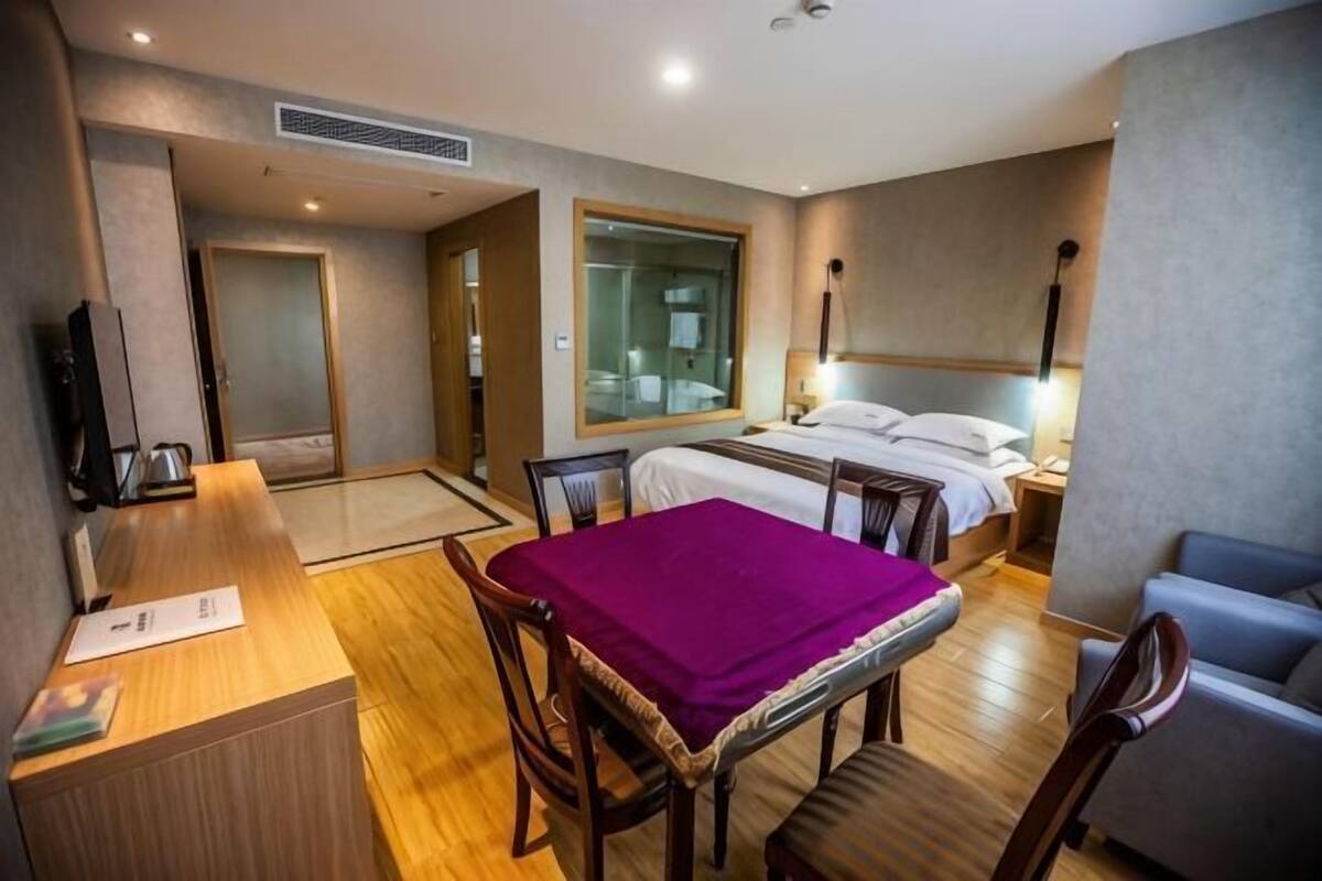 Una habitació d'hotel amb un llit, una taula de treball, una cadira, una taula amb mantell de taula morat, un sofà i una finestra gran.
