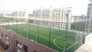 Vista de um campo esportivo com cerca, edifícios ao fundo e céu claro.