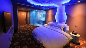 A hotel room with a bed, a nightstand, a mirror, and a bathroom visible through a glass door.