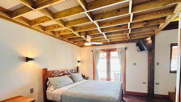 Un dormitorio con techo de madera, una cama con colcha acolchada, una mesita de noche, un televisor montado en la pared y una ventana con cortinas.