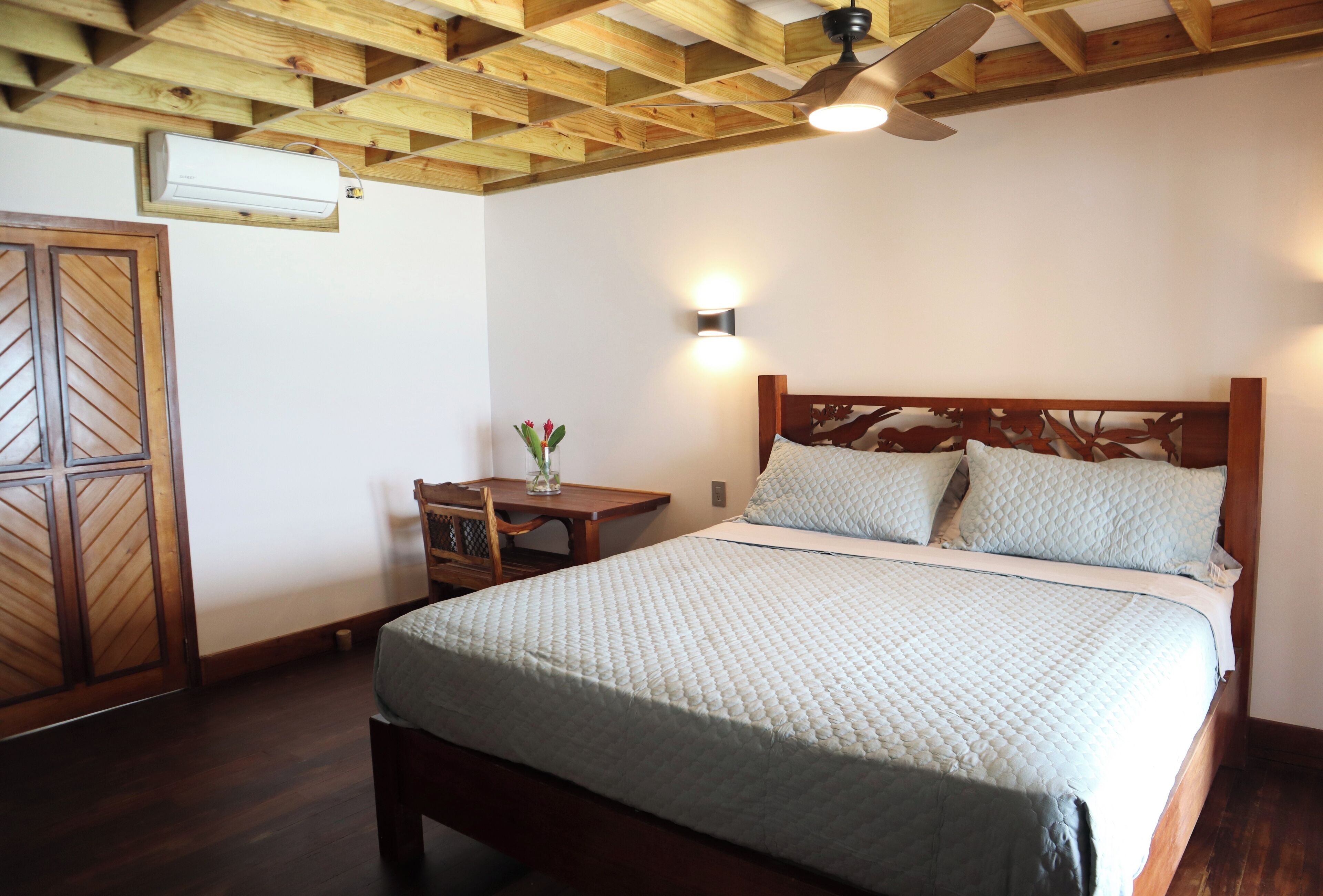 A bedroom with a wooden ceiling, a bed with a quilted bedspread, a nightstand with a vase, and a wooden wardrobe.