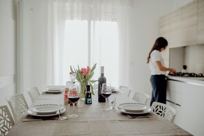 A dining table set with plates, glasses, and a bottle of wine, with a woman cooking in the kitchen.