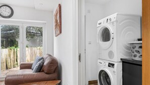 A laundry room with a washing machine and dryer, a brown leather sofa with cushions, a small wooden table, and a clock on the wall.