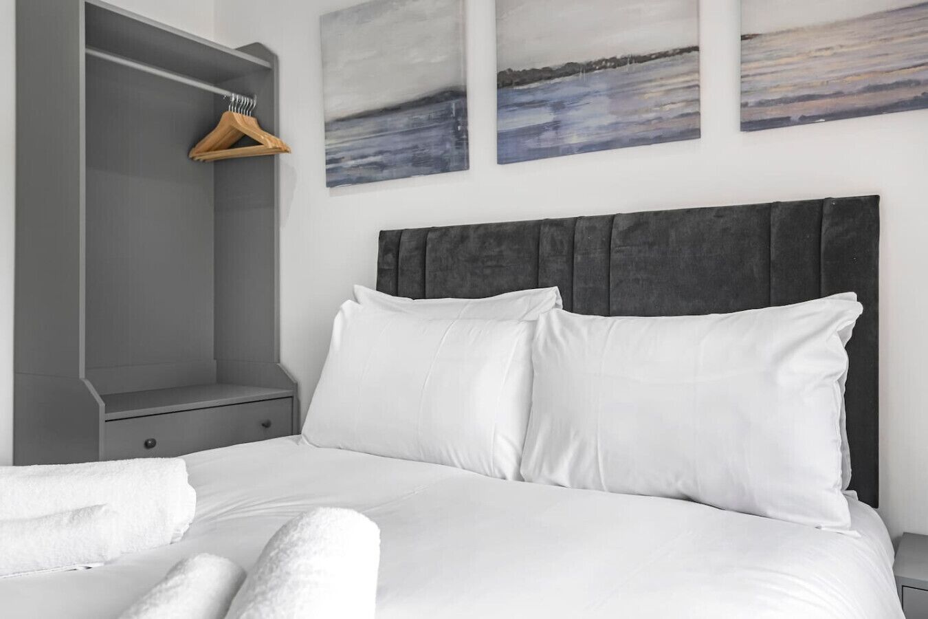 A bedroom with a bed, white pillows, a grey headboard, a wooden hanger, and three paintings on the wall.