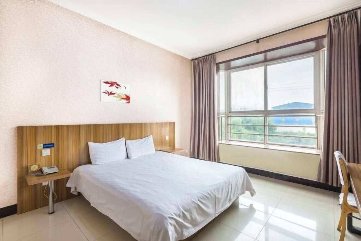 A hotel room with a bed, a nightstand, a chair, a window with curtains, and a wall-mounted artwork.