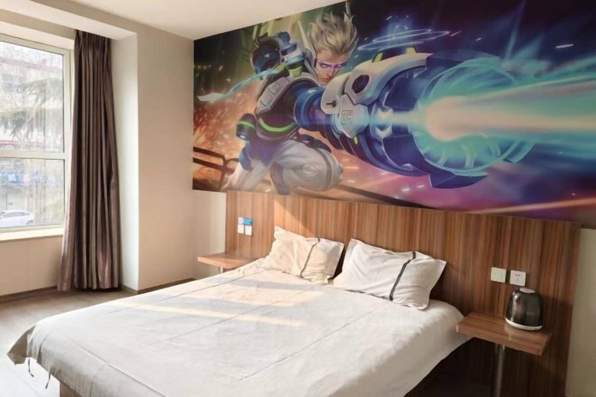 A hotel room with a large bed, a headboard with a painting, a window with curtains, and a bedside table with a lamp.