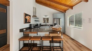A modern kitchen with a wooden ceiling, a central island with stools, and a large window with blinds. - Peaceful Sunshine Chalet with Hot Tub (Big Bear)
