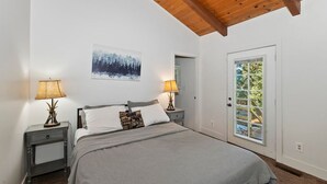 A bedroom with a bed, two bedside tables with lamps, a door with a view, and a painting on the wall. - Peaceful Sunshine Chalet with Hot Tub (Big Bear)