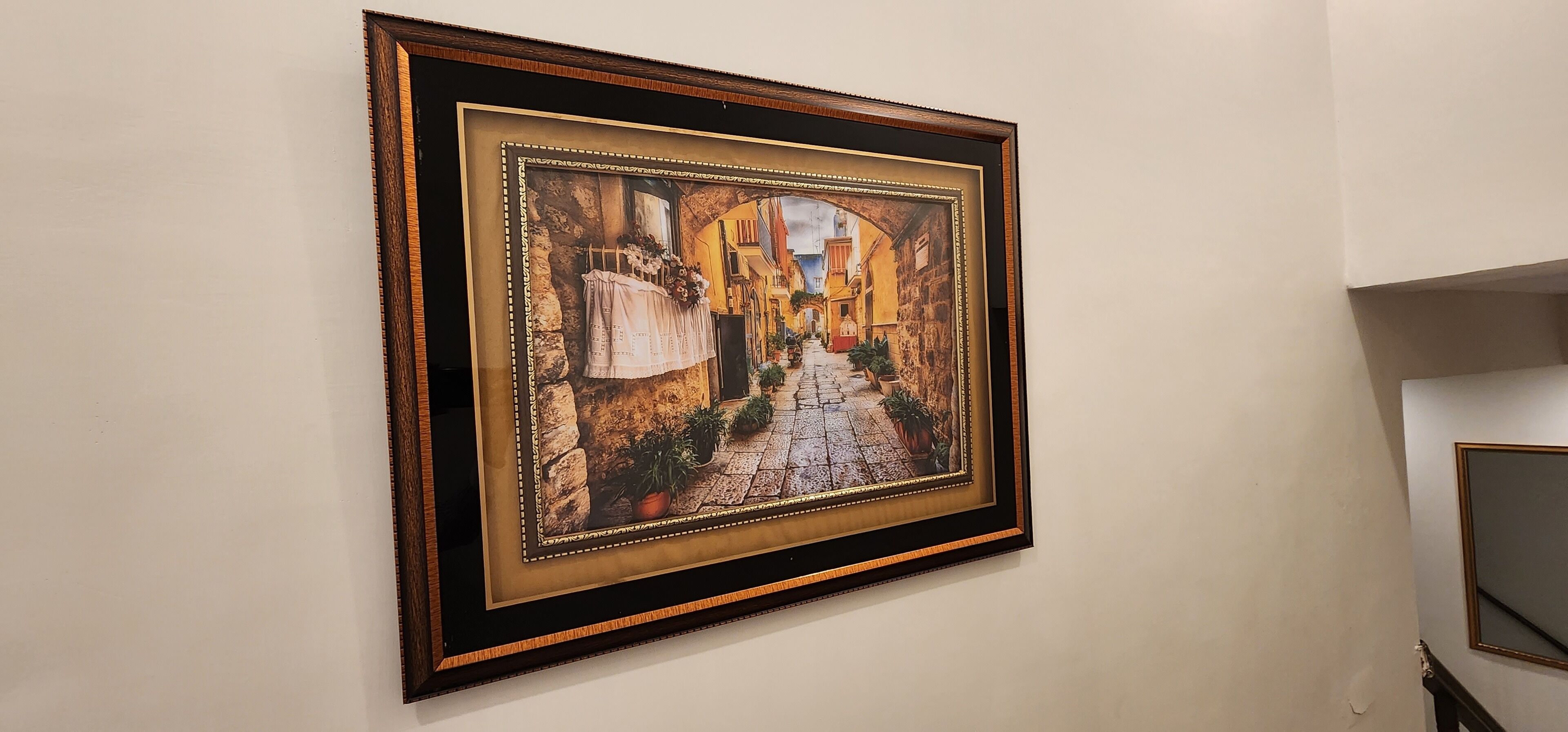 Una obra enmarcada que muestra una calle adoquinada con edificios y plantas en macetas.