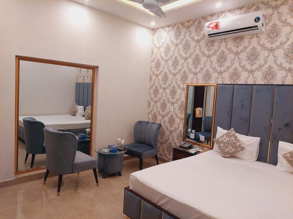 A hotel room with a large bed, a desk with a chair, a small table, a mirror, and an air conditioning unit.