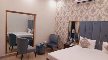 A hotel room with a large bed, a desk with a chair, a small table, a mirror, and an air conditioning unit.
