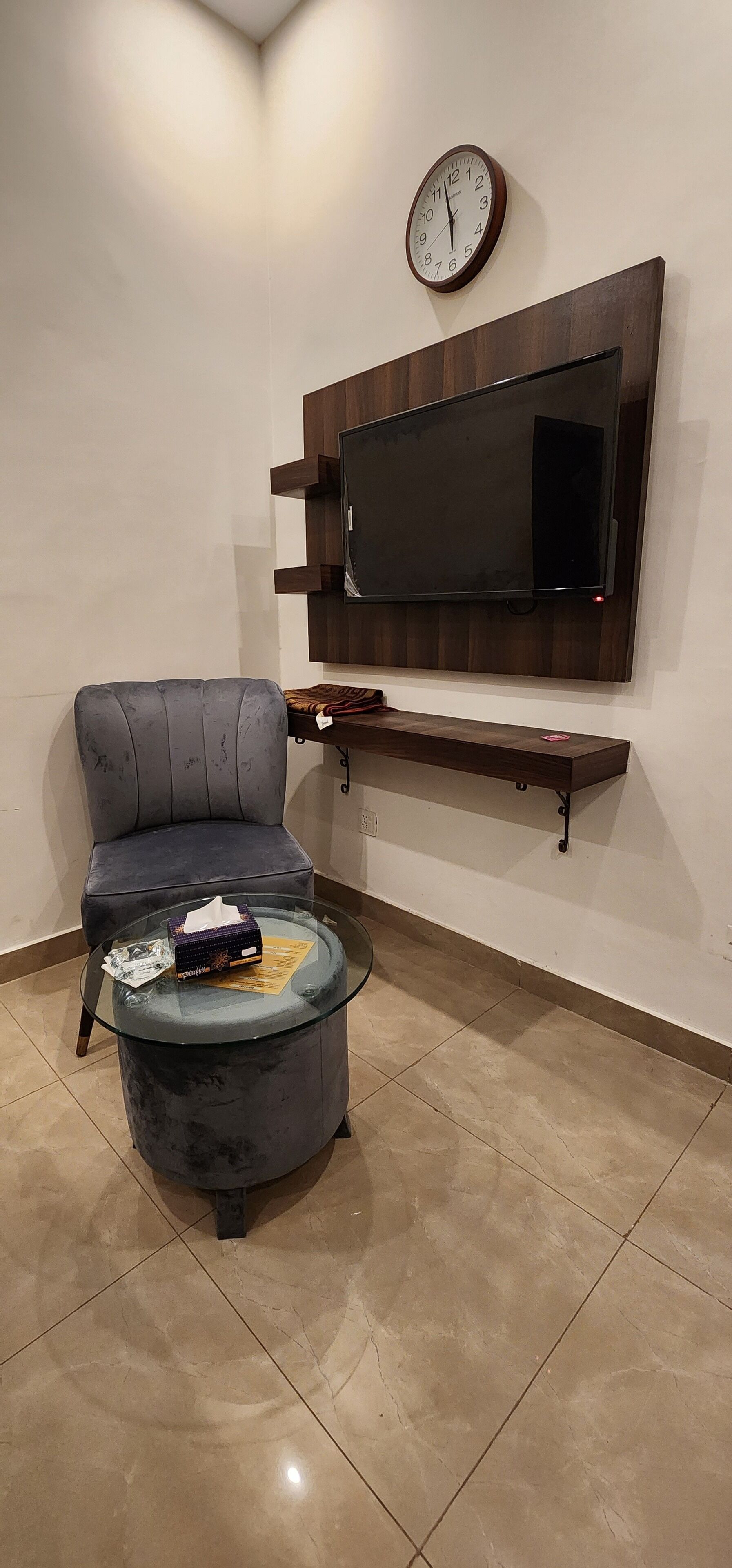 A living room with a dark grey sofa, a glass coffee table with a tissue box and a magazine, a flat-screen TV mounted on a wooden shelf, and a wall clock.