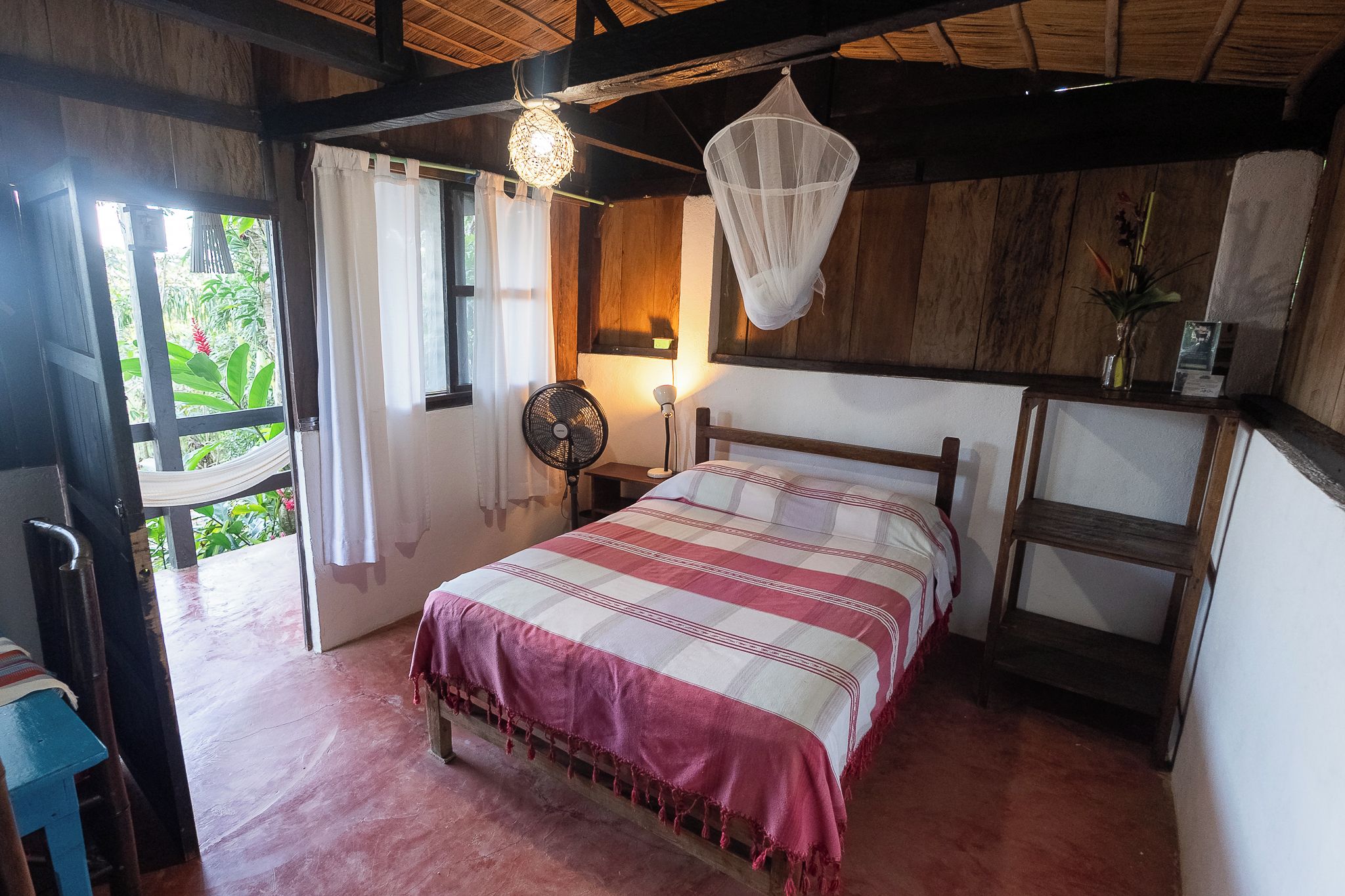 A bedroom with a bed, a fan, a nightstand, a window with curtains, and a wooden ceiling.