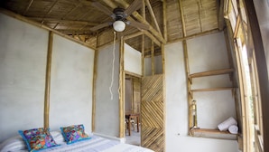 Ein Zimmer mit einem Bett, zwei bunten Kissen, einem Deckenventilator, einer Holztür und einem Regal mit Handtüchern.