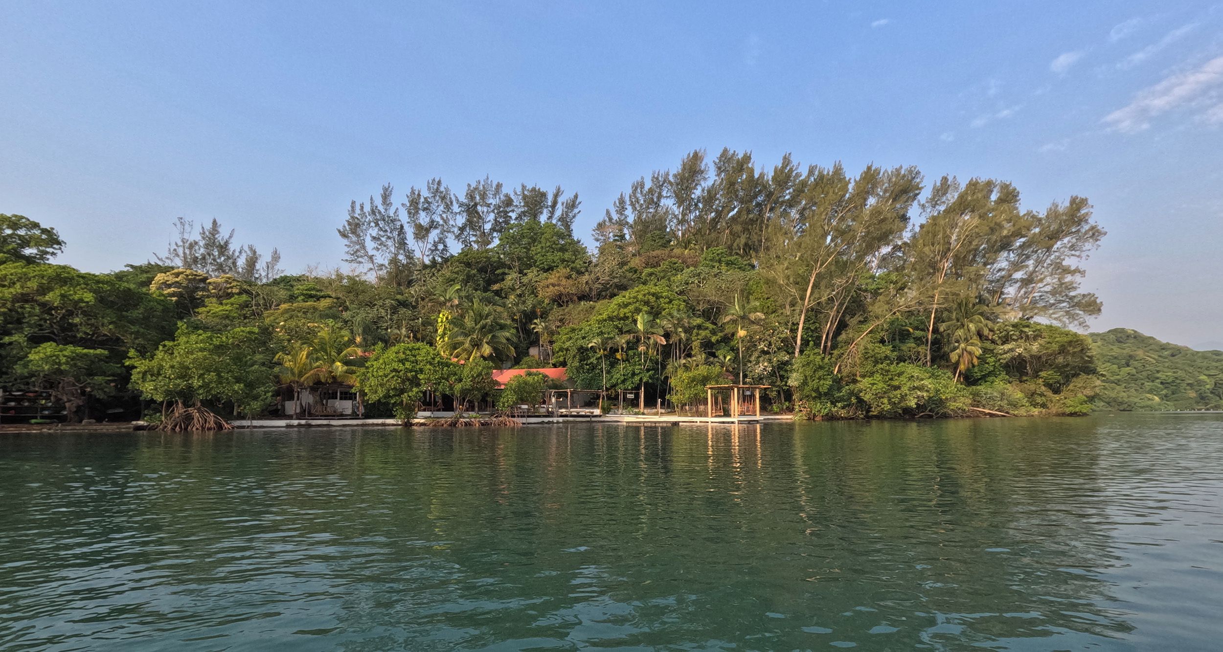 A calm lake with a dock extending into the water, surrounded by dense tropical vegetation and a clear sky.