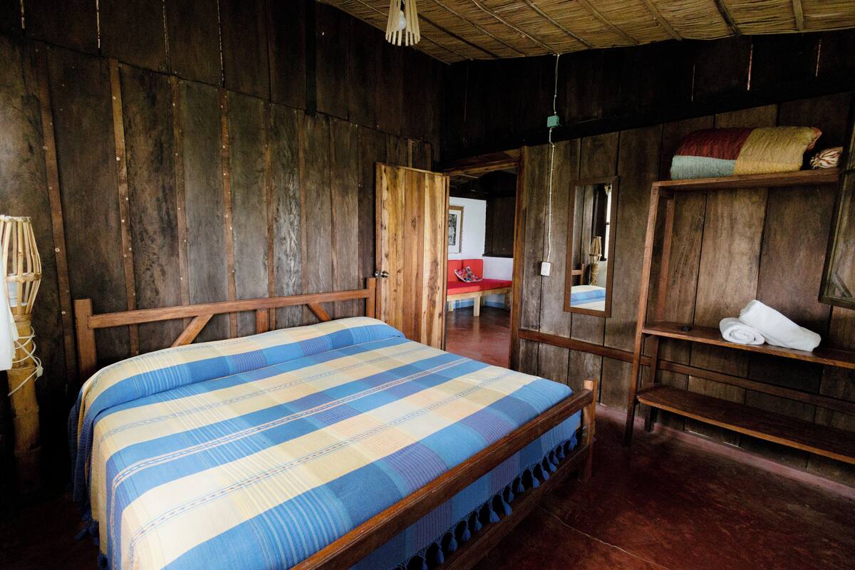 Quarto de madeira com uma cama, uma estante com toalhas e uma porta que dá para outro cômodo.