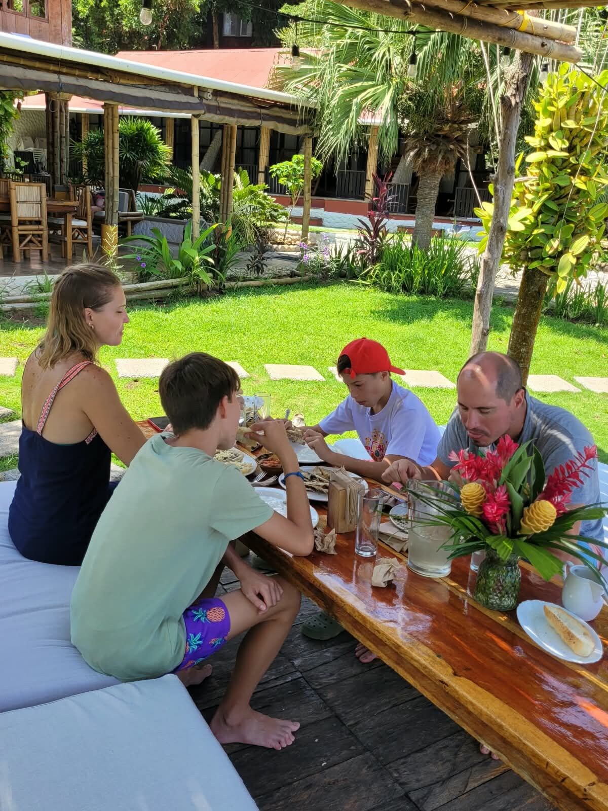 A group of people dining outdoors at a wooden table with a tropical garden backdrop.
