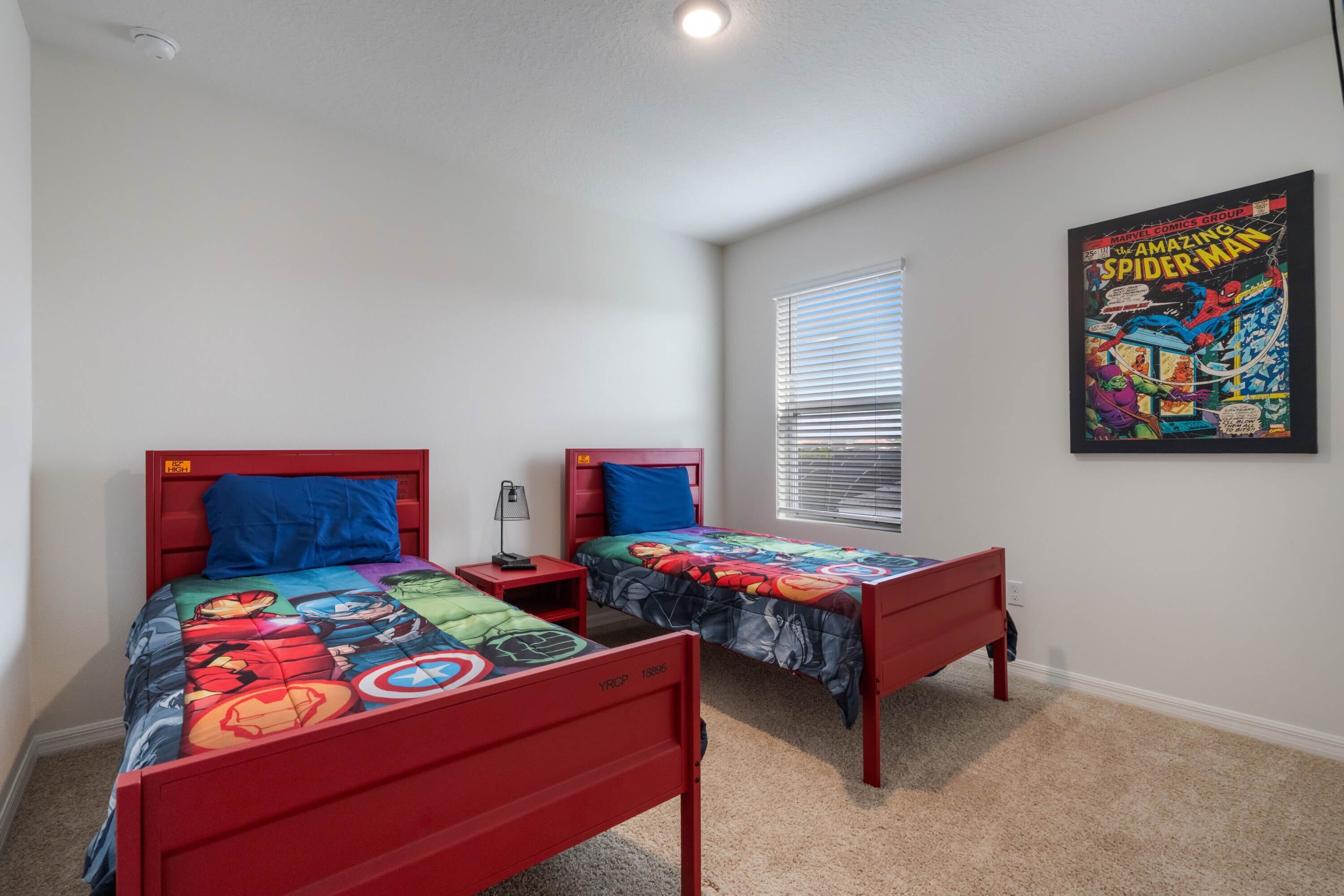 A bedroom with two red beds, each with a superhero-themed bedspread. There is a window with blinds, a wall-mounted poster of Spider-Man, and a small bedside table with a lamp.