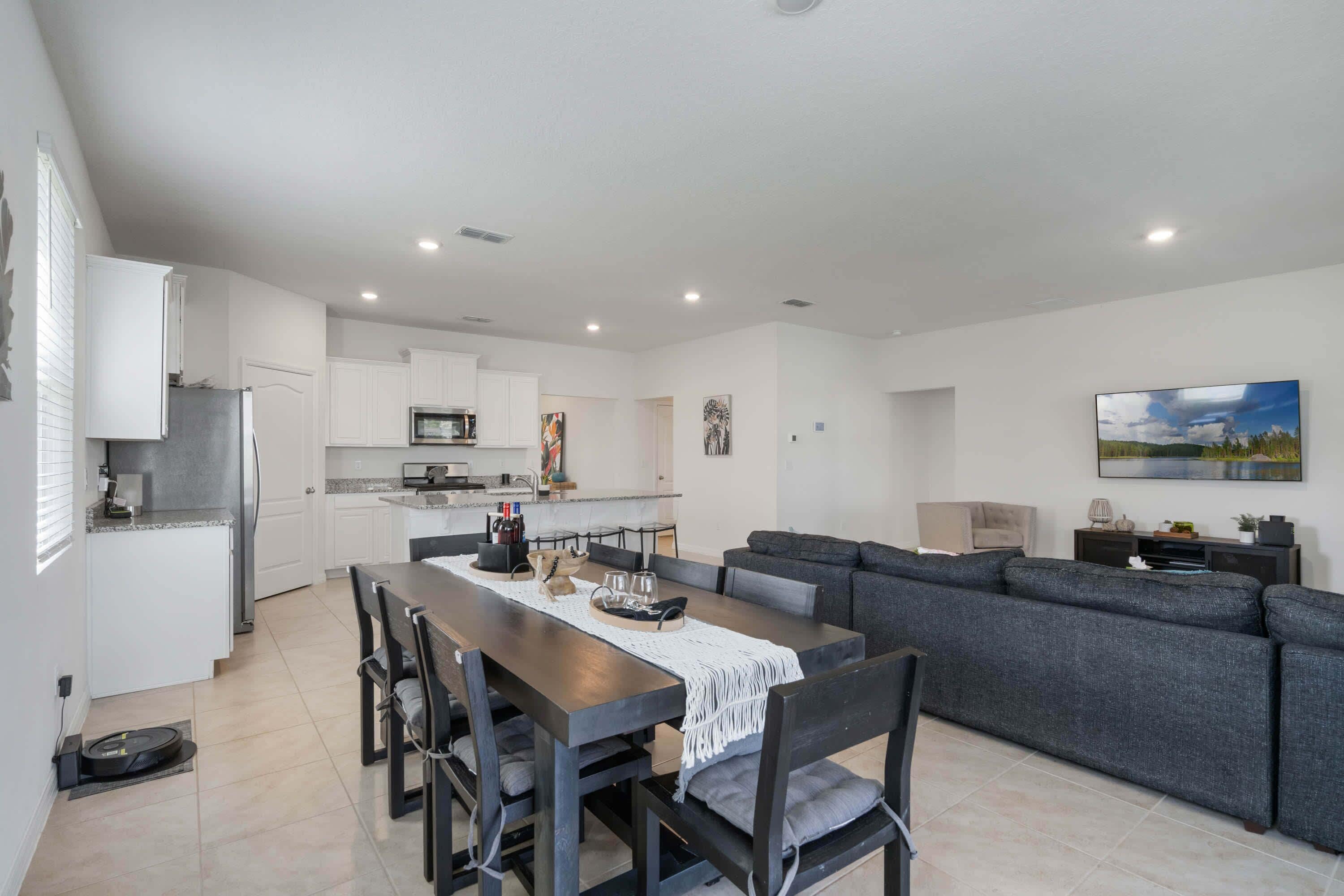 Una cocina moderna con armarios blancos, una zona de comedor con una mesa y sillas de madera oscura, y un sofá gris en la sala de estar.