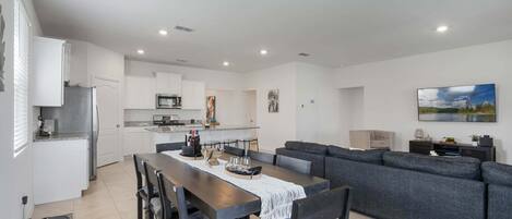 Una cocina moderna con armarios blancos, una zona de comedor con una mesa y sillas de madera oscura, y un sofá gris en la sala de estar.