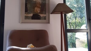 Een moderne woonkamer met een bruine bank, een lijst met een abstract schilderij, een vloerlamp en een groot raam met uitzicht op bomen.