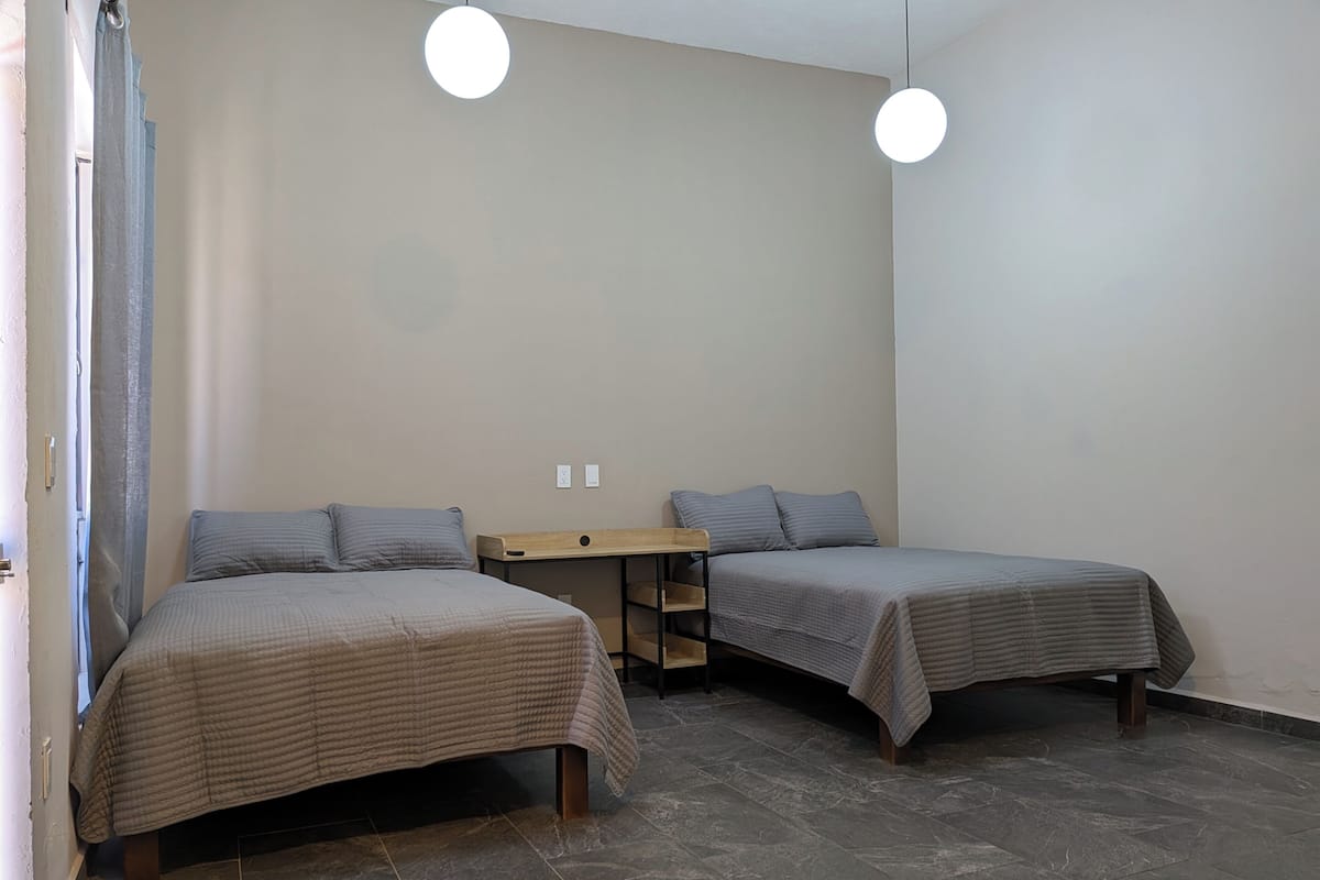 Une petite chambre propre, avec deux lits, une table de chevet en bois et deux lampes suspendues.