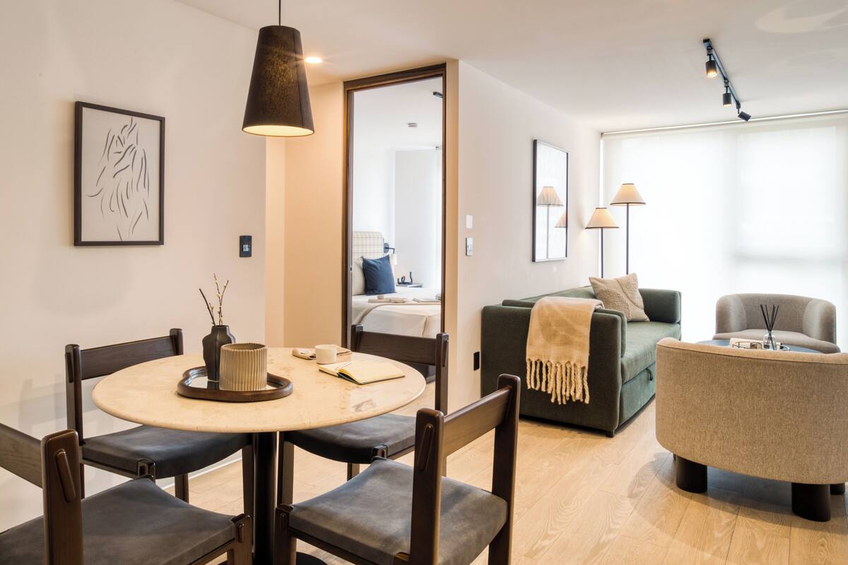 Ein modernes Wohnzimmer mit einem runden Esstisch, dunklen Holzsesseln, einem grünen Sofa und einem grauen Sessel.