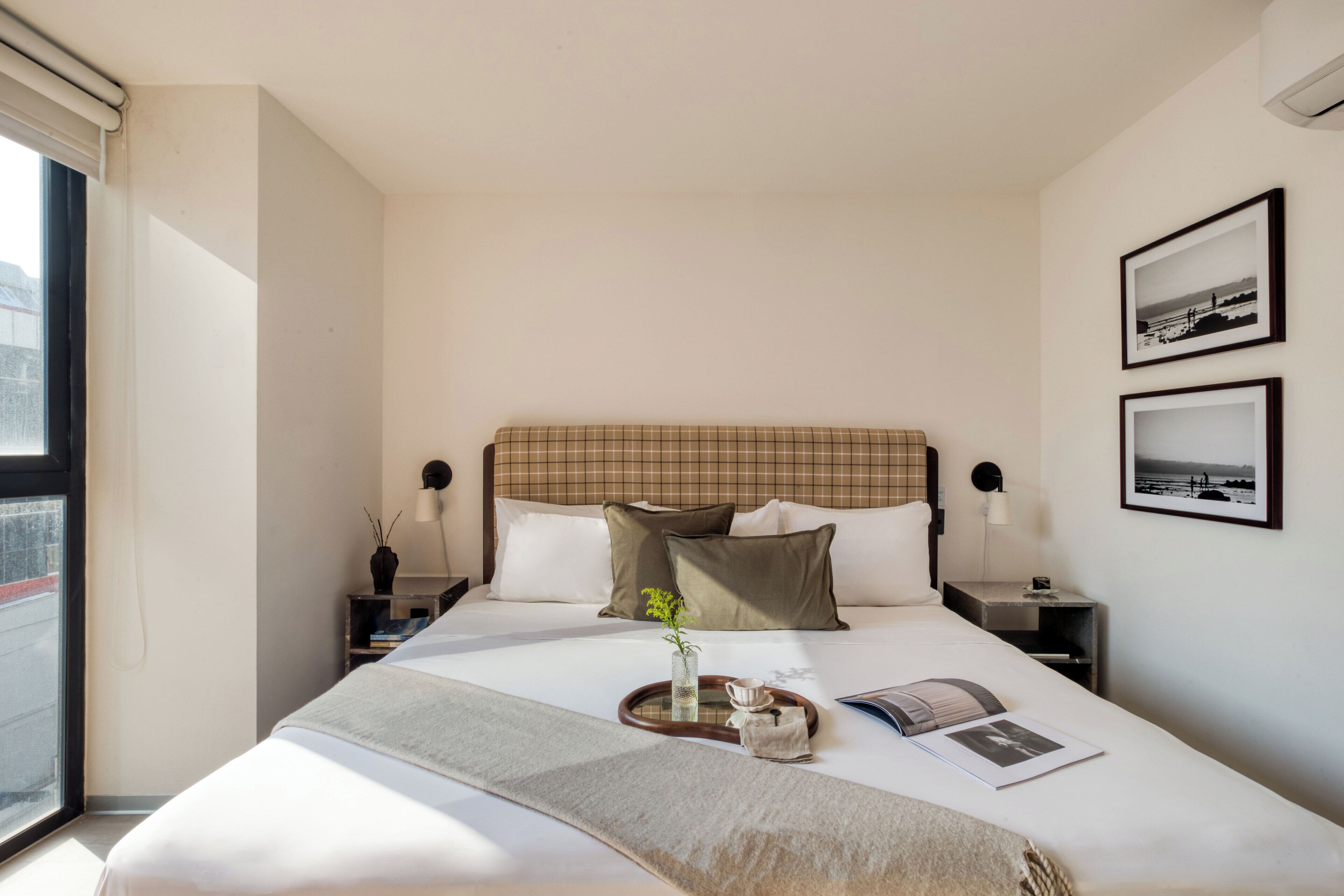 Ein ordentlich eingerichtetes Hotelzimmer mit einem großen Bett, einem Nachttisch, einem Fenster und zwei gerahmten Bildern an der Wand.