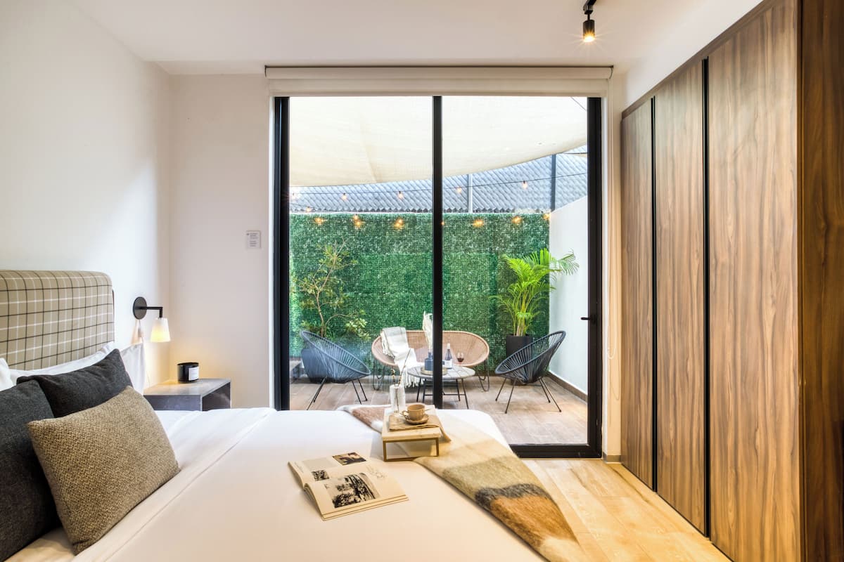 Ein modernes Schlafzimmer mit einem großen Bett, einem Nachttisch, einer Nachttruhe und Blick auf eine Außenpatio mit Essbereich und Grünflächen.