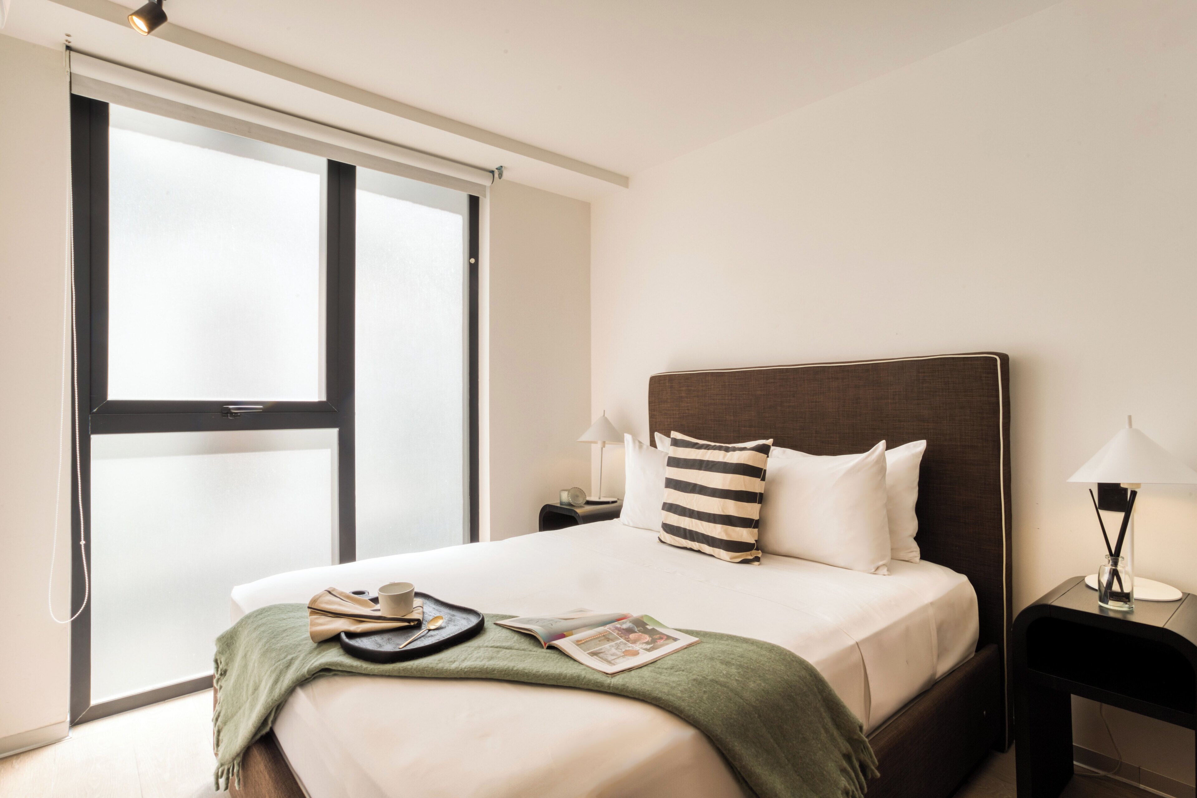 Een hotelkamer met een groot raam, een bed met een gestreept kussen, een nachtkastje met een lamp en een tijdschrift op het bed.